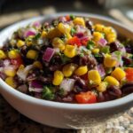 Close-up of a white bowl filled with colorful cowboy caviar featuring black beans, corn, red onion, and tomatoes.
