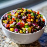 Close-up of a white bowl filled with colorful cowboy caviar, featuring black beans, corn, peppers, and red onion.