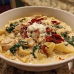 A close-up of a bowl of white chicken lasagna soup topped with ricotta, sun-dried tomatoes, and spinach.