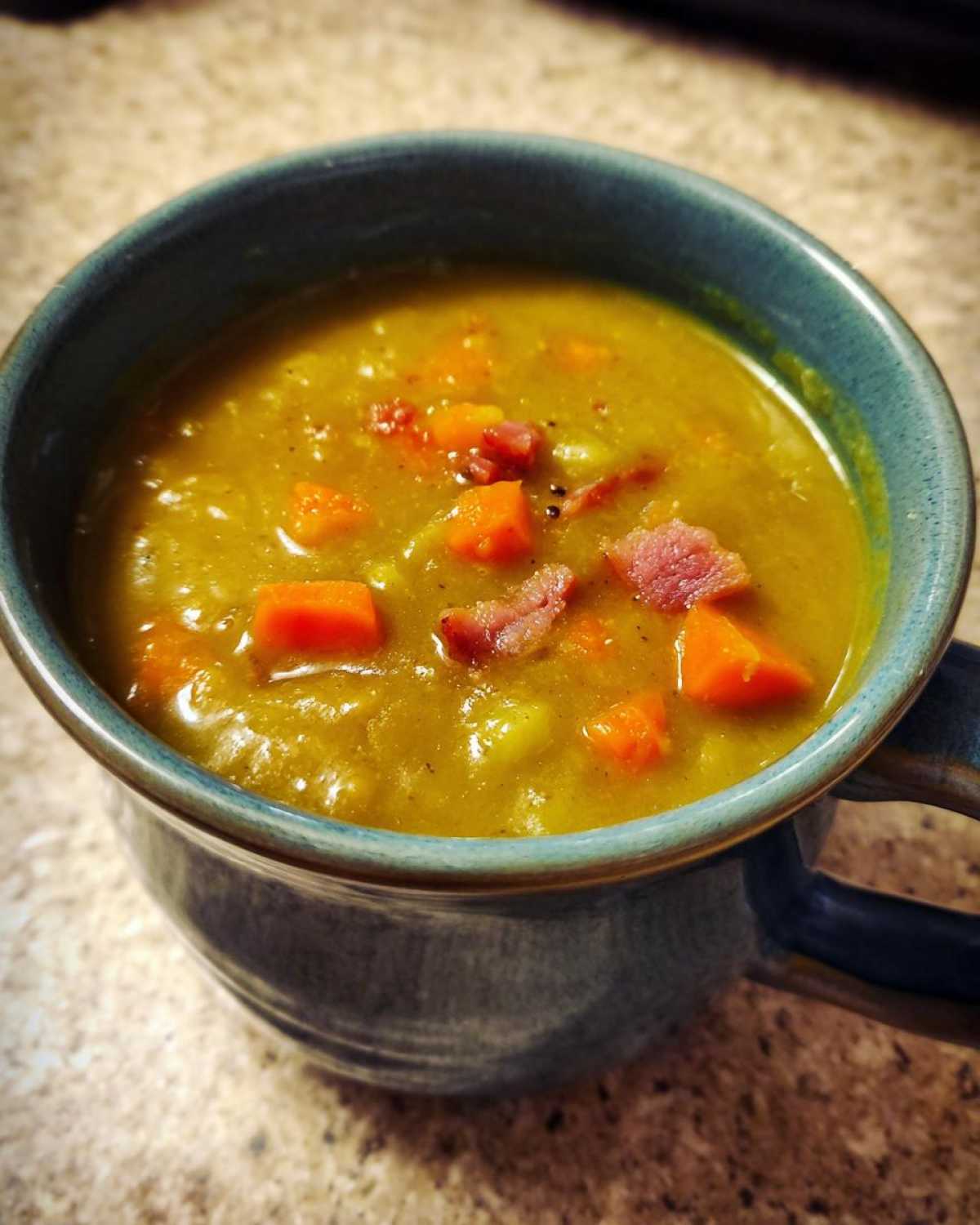 Close-up of a thick, yellow pea soup served in a blue mug, topped with diced carrots and pieces of ham.