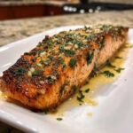 A close-up of a perfectly cooked air fryer salmon fillet topped with minced garlic and fresh parsley, resting in butter sauce.
