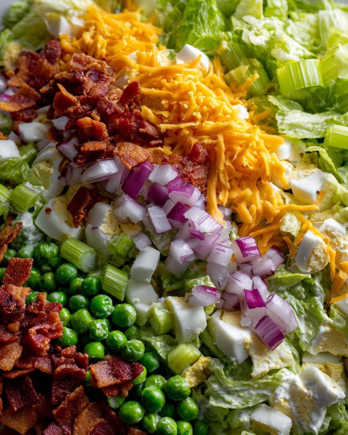 Close-up of layered ingredients for 7 layer salad including lettuce, peas, bacon, cheese, and red onion.