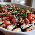 Close-up of a hot, baked bruschetta dip topped with diced tomatoes, fresh basil, and a balsamic glaze drizzle.