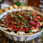 Close-up of a hot, baked bruschetta dip topped with diced tomatoes, fresh basil, and a balsamic glaze drizzle.