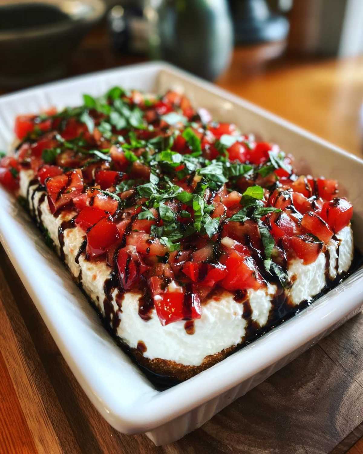 A creamy, layered bruschetta dip topped with diced tomatoes, fresh basil, and balsamic glaze in a white rectangular dish.