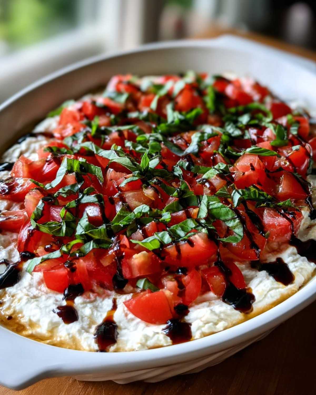 Close-up of a creamy, layered bruschetta dip topped with diced tomatoes, fresh basil, and balsamic glaze.