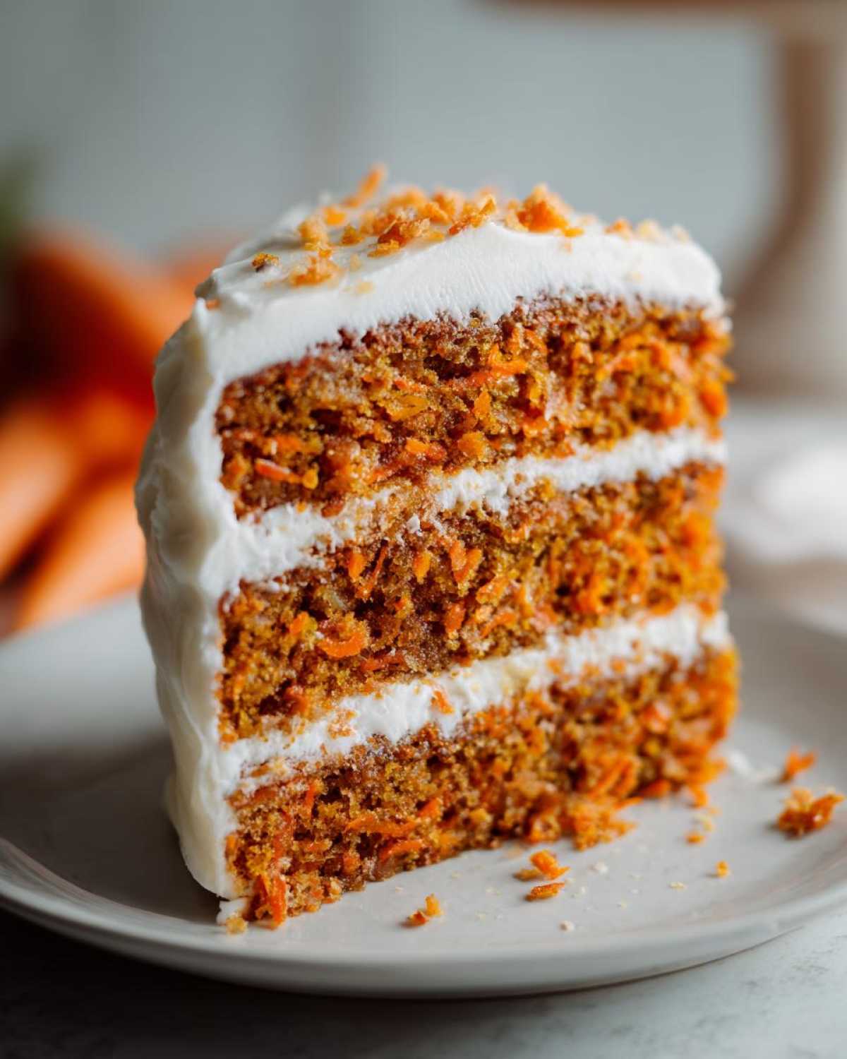 A tall, moist slice of carrot cake recipe with three layers, cream cheese frosting, and carrot shavings on top.