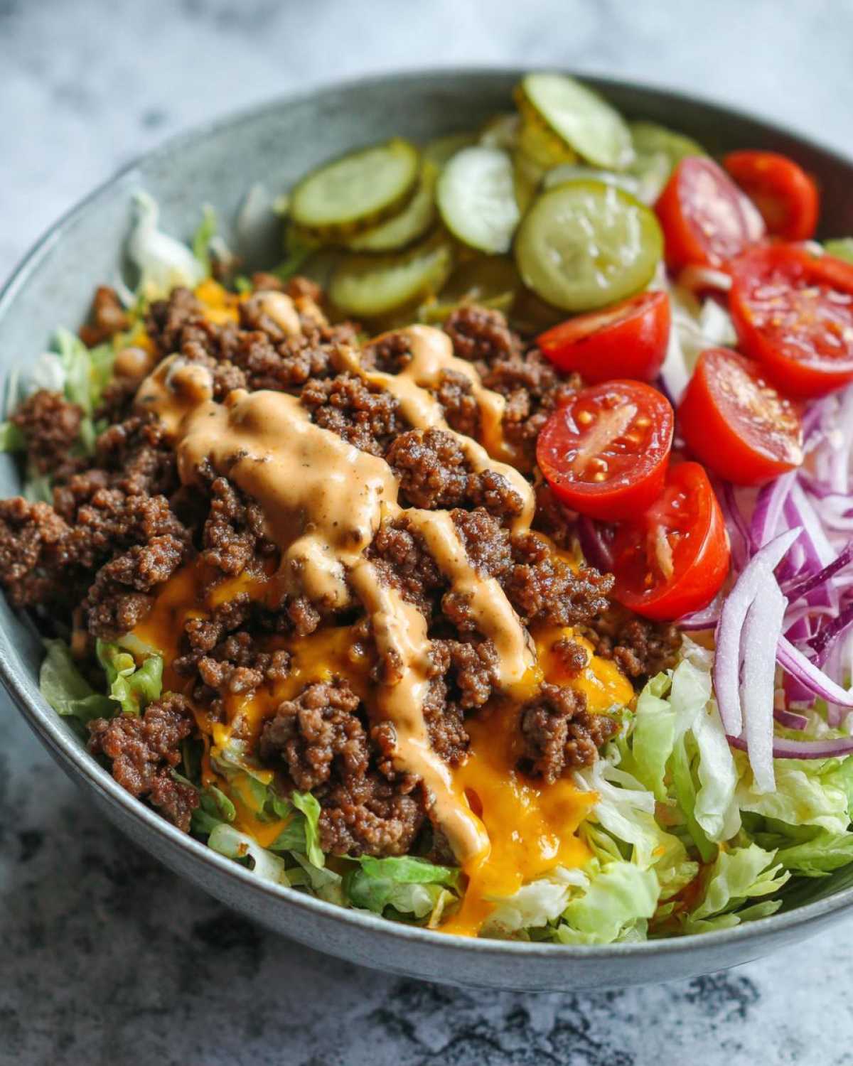 A close-up of a vibrant cheeseburger bowl featuring seasoned ground beef, melted cheese, special sauce, lettuce, pickles, tomatoes, and red onion.