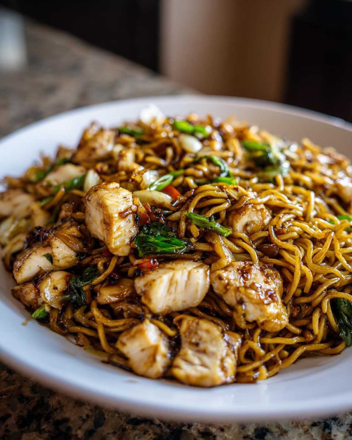 A close-up shot of a white plate filled with glossy, saucy chicken chow mein recipe noodles and chunks of white chicken meat.