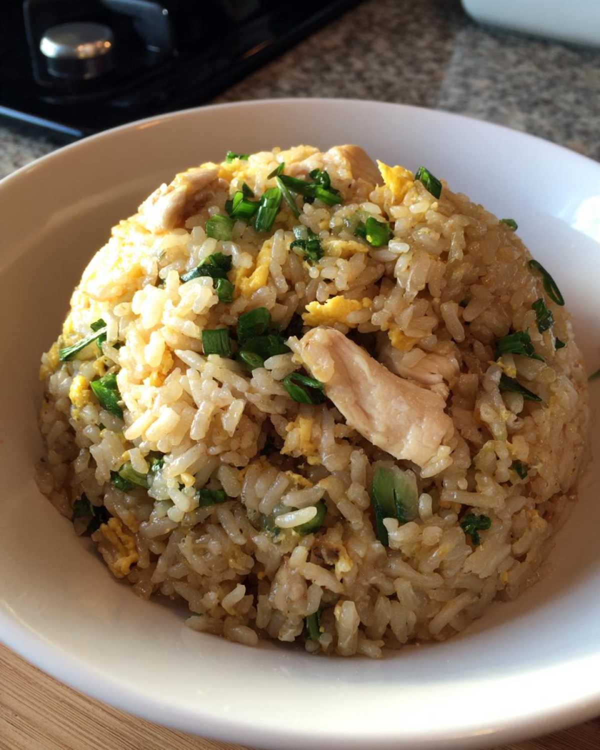 A close-up of a white bowl filled high with delicious chicken fried rice, topped with pieces of chicken and green onions.