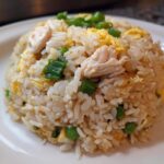 A mound of homemade chicken fried rice with visible pieces of chicken, scrambled egg, and green onions on a white plate.