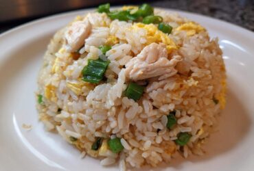 A mound of homemade chicken fried rice with visible pieces of chicken, scrambled egg, and green onions on a white plate.