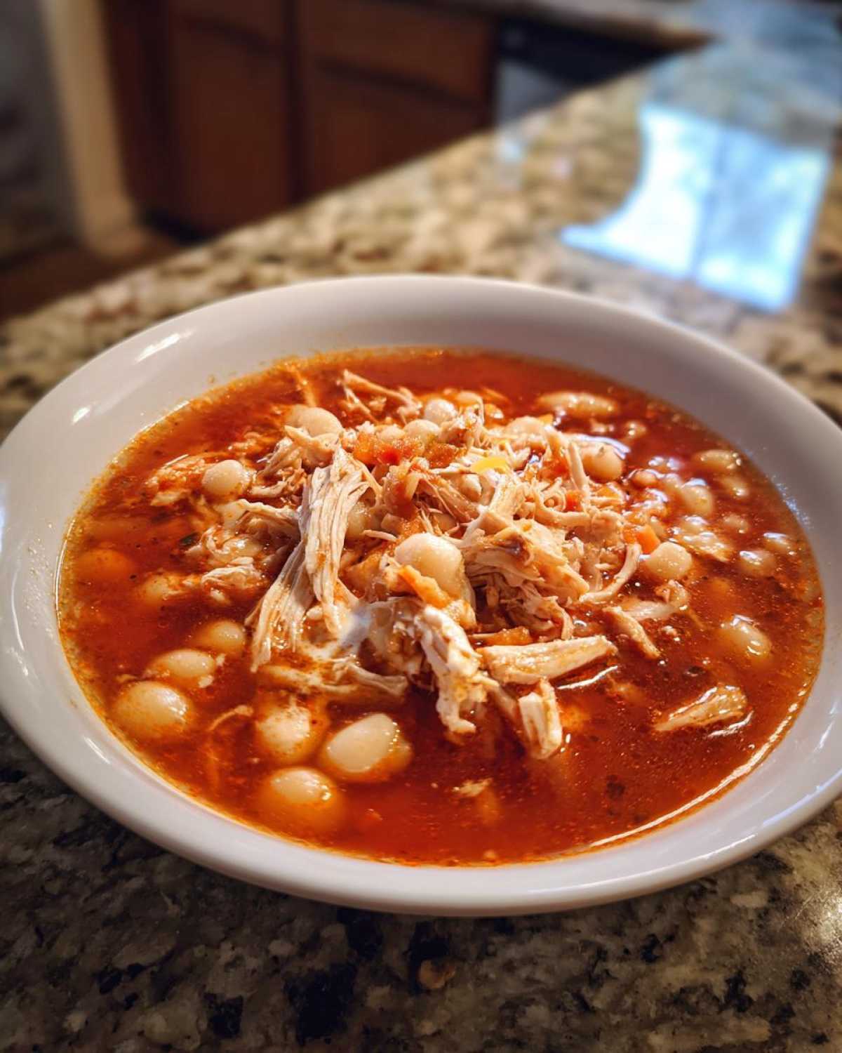 A close-up of a white bowl filled with rich red broth, shredded chicken, and hominy for amazing chicken pozole.