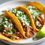 Close-up of three flavorful chicken tacos filled with shredded meat, topped with white cheese, onions, and cilantro, served with a lime wedge.