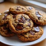 A stack of freshly baked, thick chocolate chip cookies with melted chocolate pools, served on a white plate.