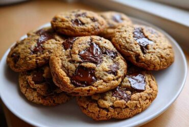 A stack of freshly baked, thick chocolate chip cookies with melted chocolate pools, served on a white plate.