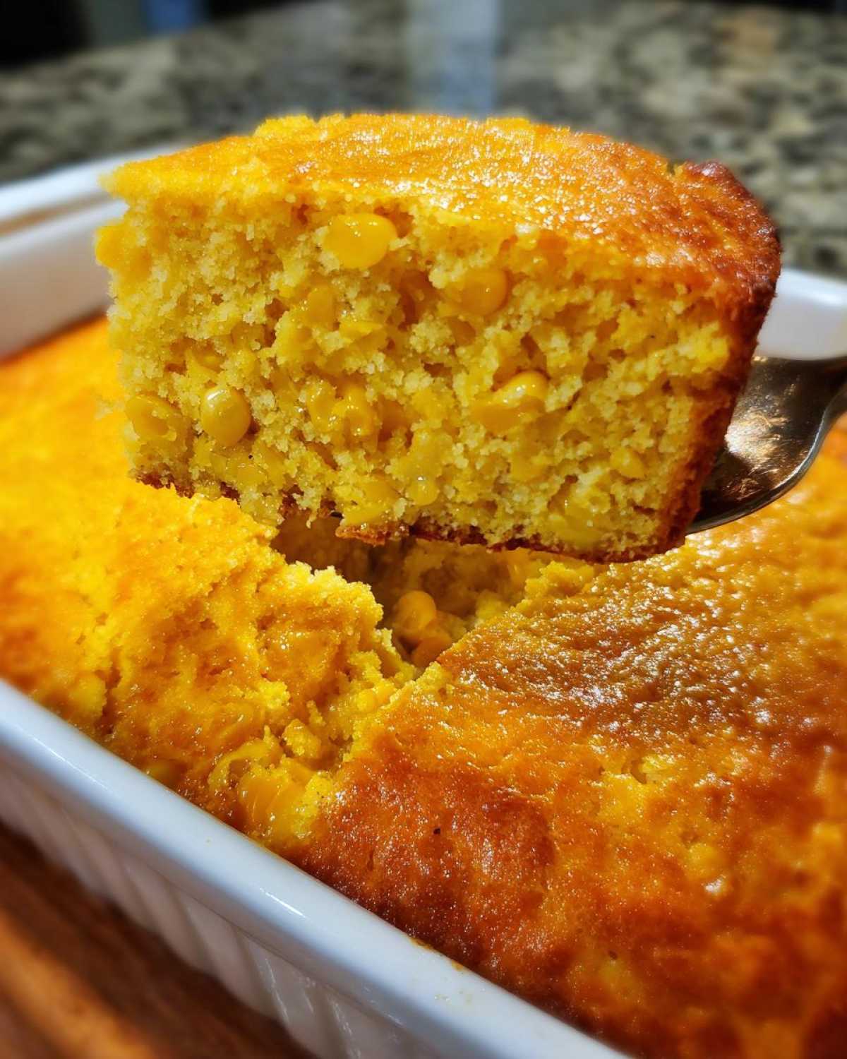 A serving spoon lifts a moist, golden slice of cornbread casserole, rich with whole corn kernels, from a white baking dish.