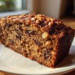 Close-up of a moist slice of date nut bread topped with walnuts, sitting on a white plate.