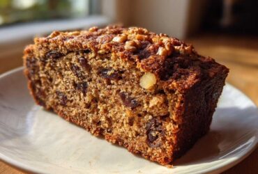 Close-up of a moist slice of date nut bread topped with walnuts, sitting on a white plate.