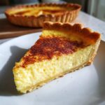 Close-up of a thick slice of creamy egg custard tart dusted with cinnamon on a white plate.