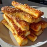 A close-up of a tall stack of golden brown garlic breadsticks topped with melted cheese and herbs.