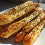 A close-up of several golden brown, herb-topped garlic breadsticks stacked on a white plate.