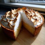 A slice cut from a whole lemon meringue pie showing the thick, toasted meringue topping and bright yellow filling.