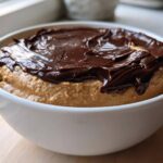 Close-up of a white bowl filled with creamy peanut butter dip topped with a layer of melted chocolate.