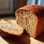 A close-up of a freshly baked whole wheat bread loaf, with one slice cut and resting against it on a wooden cutting board.