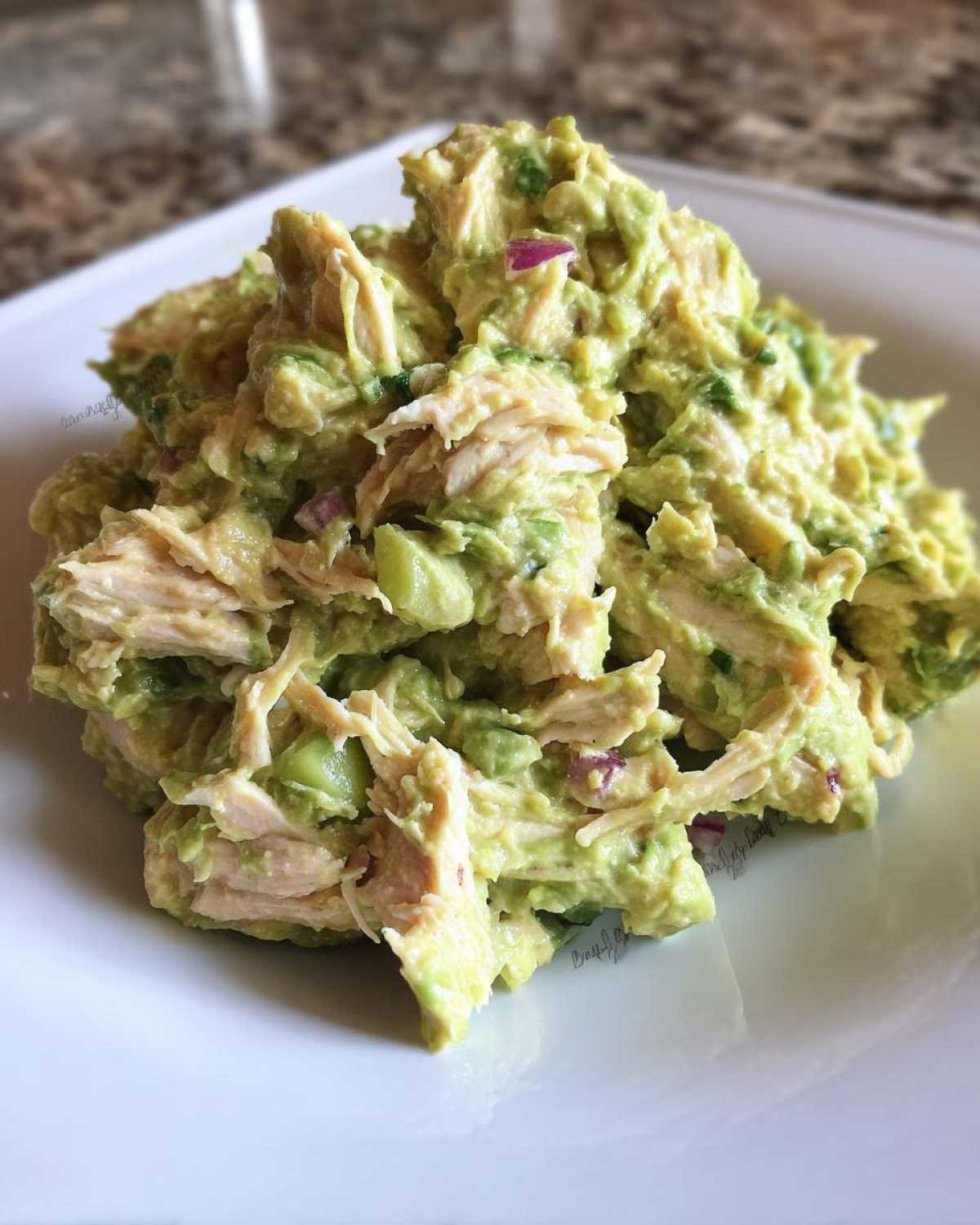 A mound of creamy avocado chicken salad mixed with shredded chicken and diced red onion served on a white plate.