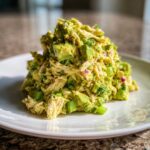 A mound of creamy avocado chicken salad featuring shredded chicken, chunks of avocado, and green onions on a white plate.