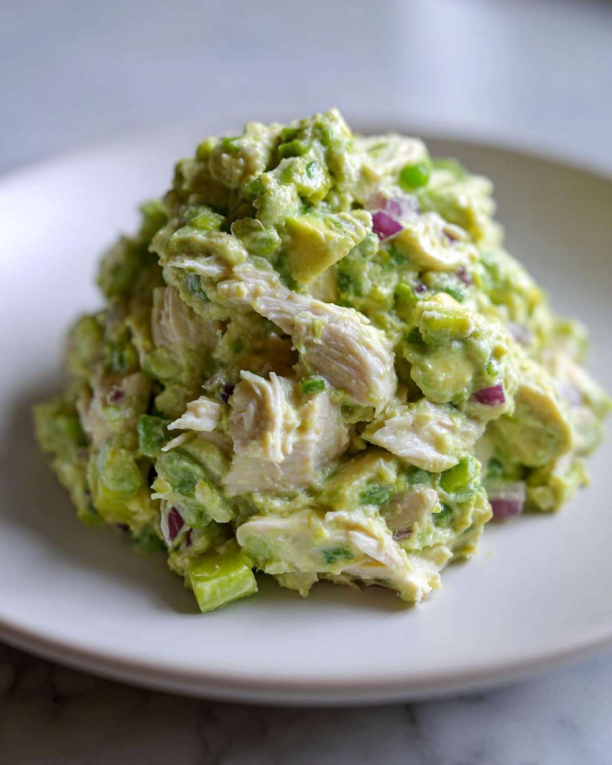 A mound of creamy avocado chicken salad featuring shredded chicken, chunks of avocado, and red onion on a white plate.