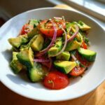Close-up of a fresh avocado salad with chunks of avocado, cherry tomatoes, red onion, and herbs in a white bowl.