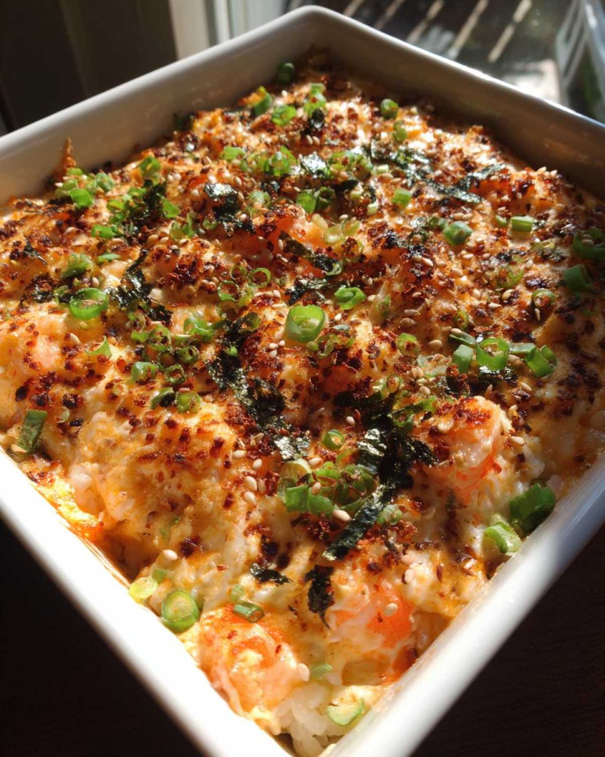 Close-up of a freshly baked sushi bake in a white dish, topped with creamy sauce, shrimp, scallions, and sesame seeds.