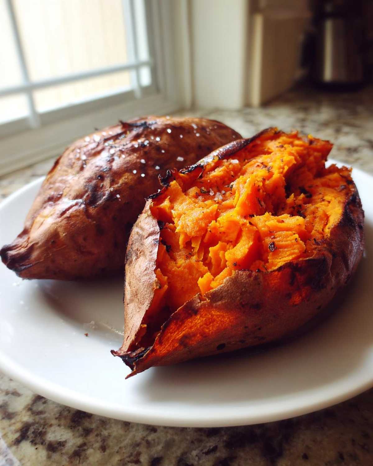 Two perfectly baked sweet potatoes on a white plate, one split open showing fluffy orange interior seasoned with salt.