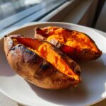 Two perfectly baked sweet potatoes, split open to reveal bright orange, fluffy flesh, sitting on a white plate.