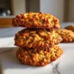 A vertical stack of three golden-brown banana oatmeal cookies resting on a white marble surface.