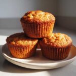 Three golden-brown banana protein muffins stacked on a light ceramic plate, captured in dramatic sunlight.