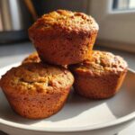A stack of three golden-brown banana protein muffins sitting on a light-colored plate, captured in bright sunlight.