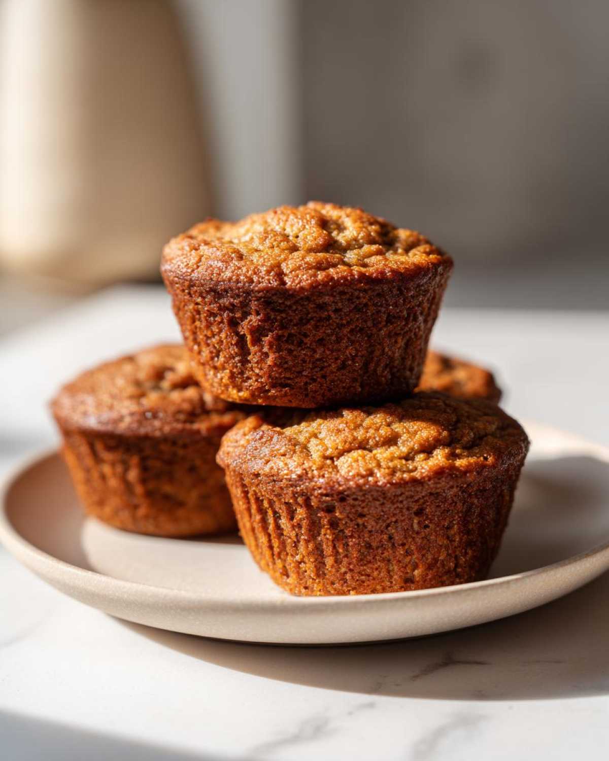 A stack of four golden brown banana protein muffins resting on a light ceramic plate.