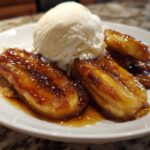 Close-up of caramelized bananas foster halves topped with a scoop of vanilla ice cream on a white plate.