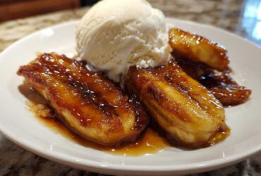 Close-up of caramelized bananas foster halves topped with a scoop of vanilla ice cream on a white plate.