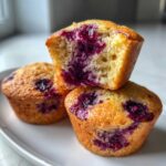 Three golden-brown berry-filled protein muffins stacked on a white plate, one cut in half showing the moist interior.
