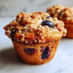 A golden brown blueberry muffin with a generous, sugary crumb topping sits on a marble surface.