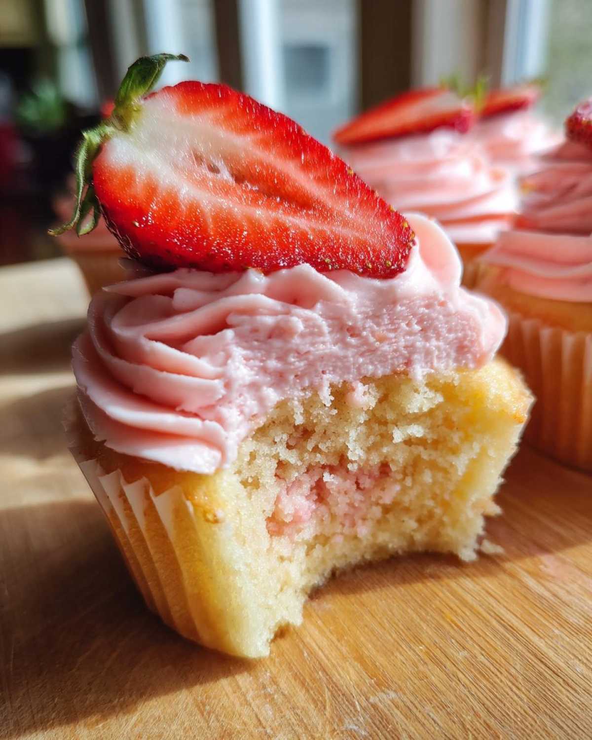 Close-up of a bitten strawberry cupcake showing the moist cake, pink filling, and strawberry frosting topped with a fresh strawberry slice.