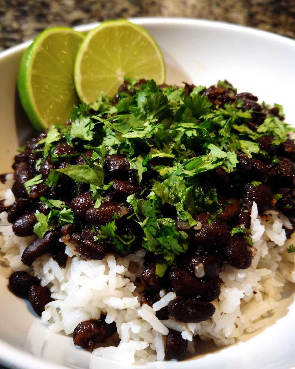 Close-up of a bowl of white rice topped with seasoned black beans and fresh cilantro, garnished with lime wedges.