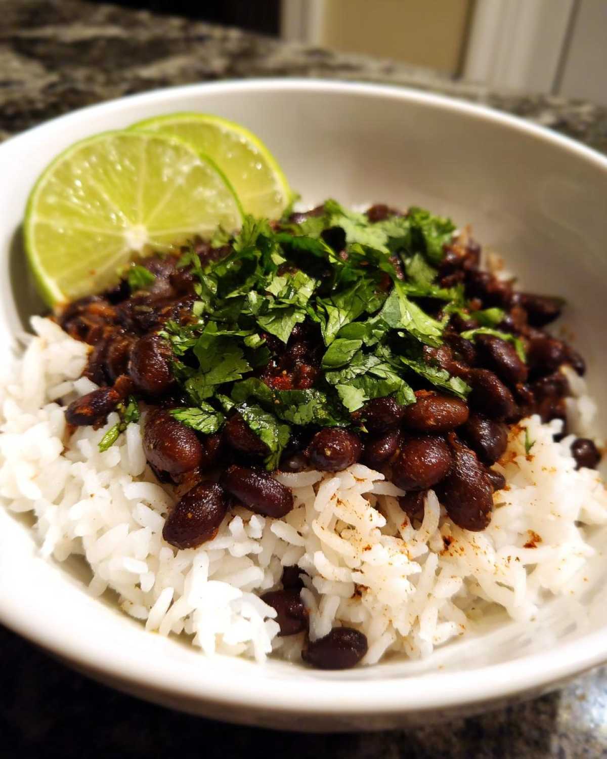A white bowl filled with white rice topped with seasoned black beans and fresh cilantro, garnished with lime slices.