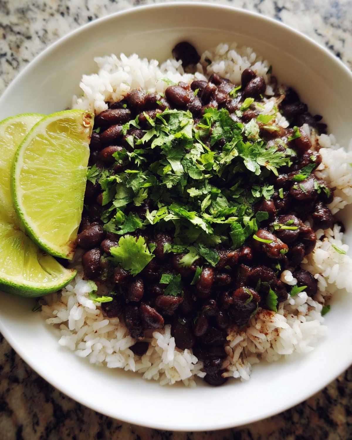 A white bowl filled with fluffy white rice topped generously with seasoned black beans and fresh cilantro, served with lime wedges.