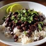 A close-up of a bowl of white rice topped with seasoned black beans and fresh cilantro, garnished with lime wedges.