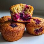 A stack of golden brown blueberry protein muffins, with the top one cut in half showing the moist interior.